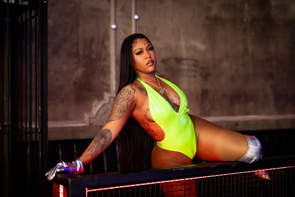 Tattooed woman in neon yellow bodysuit posing athletically on stage railing under dramatic lighting.