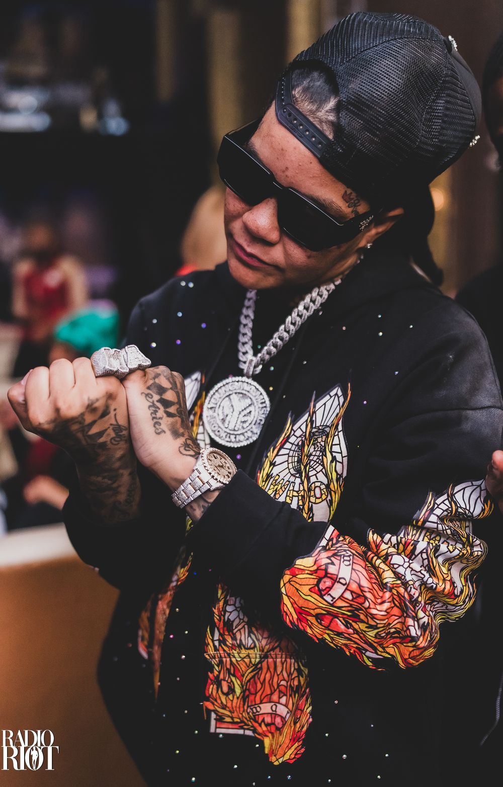 Male rapper wearing sunglasses and diamond chain necklace with tattooed hands at a nightclub event.