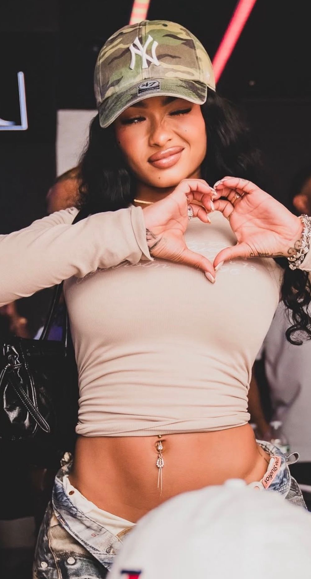 Young woman in camo NY Yankees cap making heart shape with hands at nightclub event.
