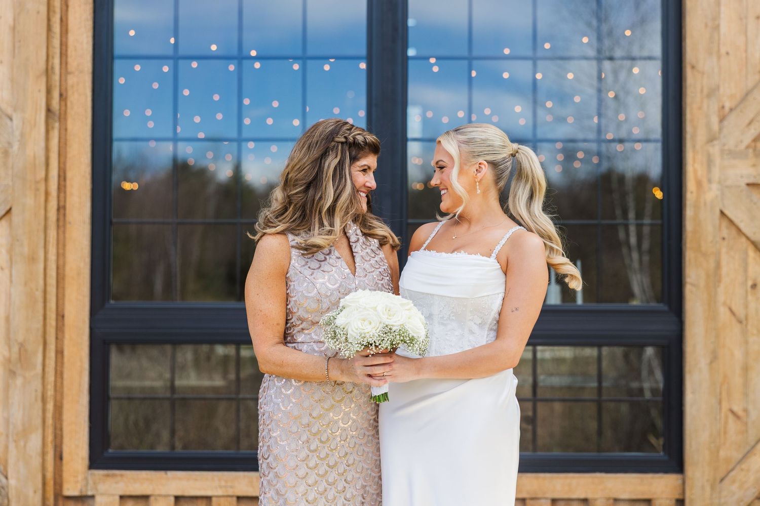 Two brides smiling at each other, one holding a white bouquet, posing in front of a window with fairy lights.