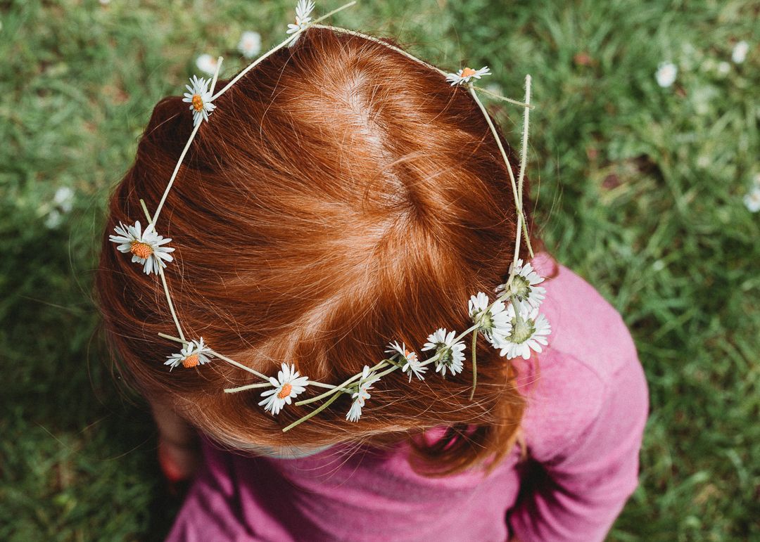 Red-haired girl wearing a delicate daisy flower crown, viewed from above, sitting on green grass.