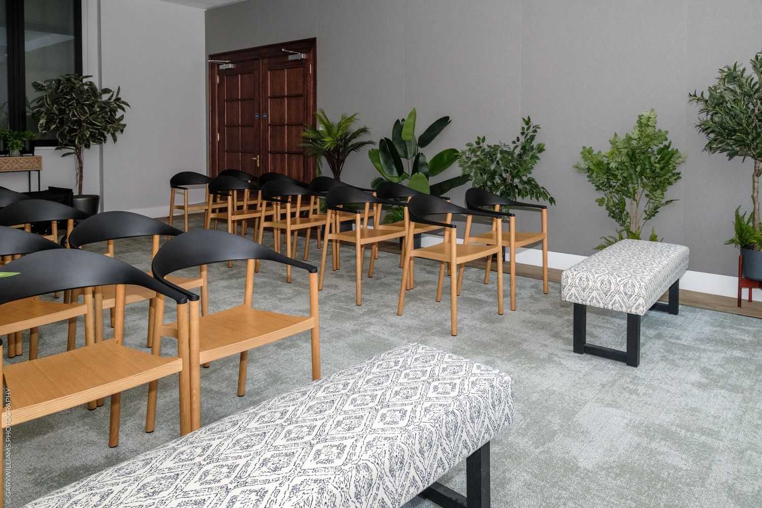 Modern waiting room with wooden chairs, black seats, patterned benches, and tropical plants on gray tile floor.