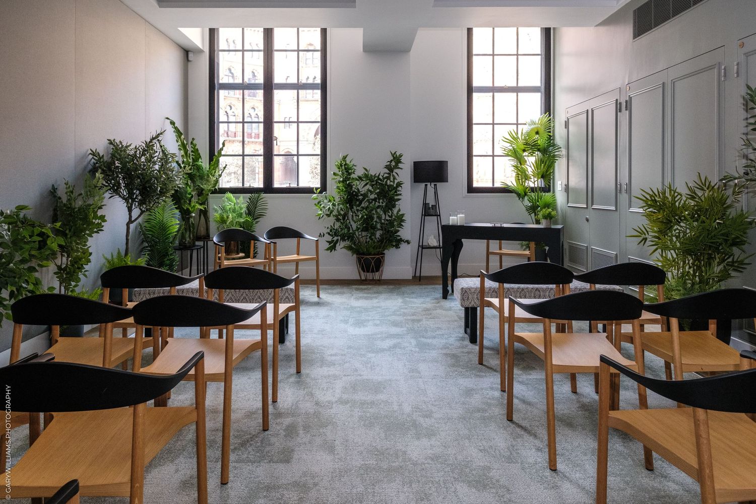 Elegant event space with wooden chairs arranged in rows, lush green plants, and large windows in a minimalist white room.