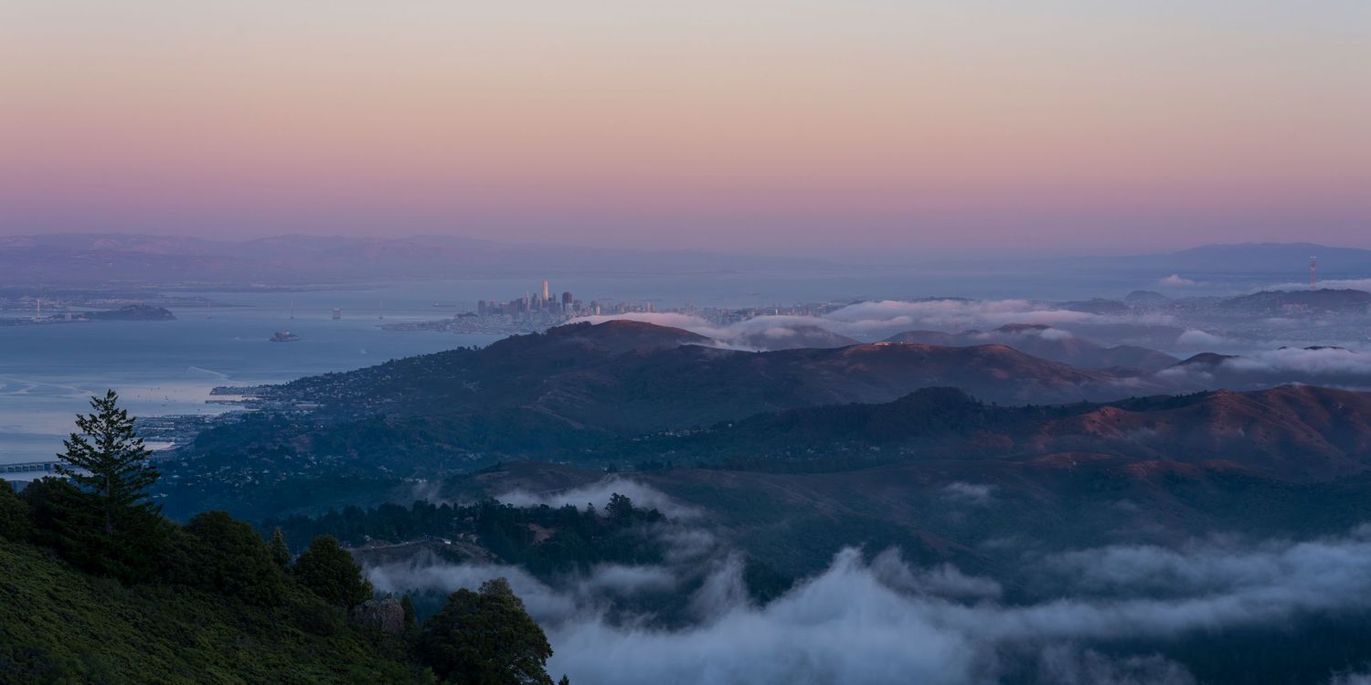Panoramic sunset view of San Francisco skyline from misty hillside with fog rolling through valleys at dusk.