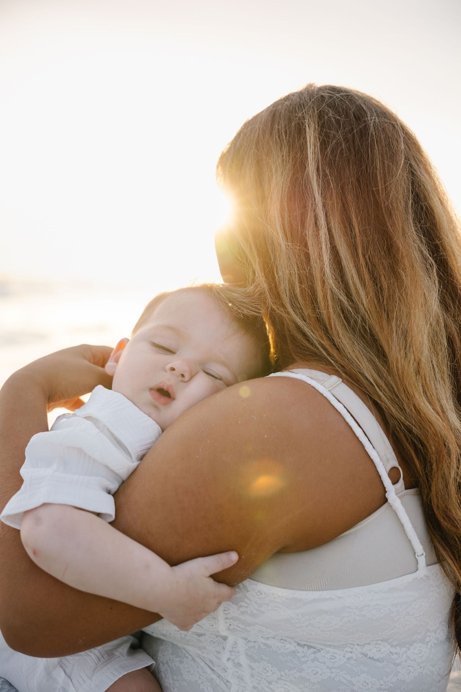 A mother holds her sleeping baby against her shoulder on a sunny beach, bathed in warm golden backlight.