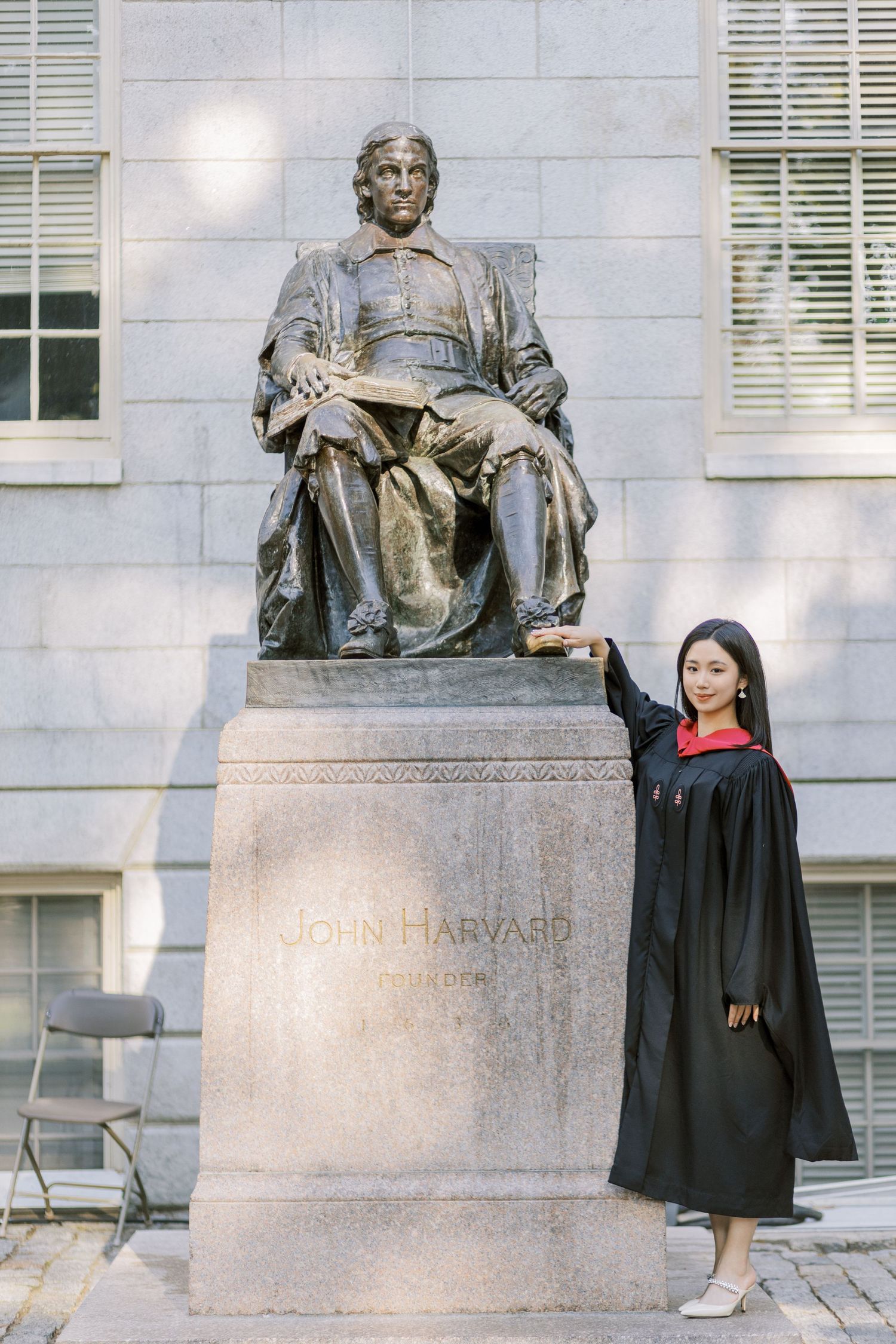 Harvard graduate in cap and gown poses proudly beside the iconic John Harvard statue on campus.