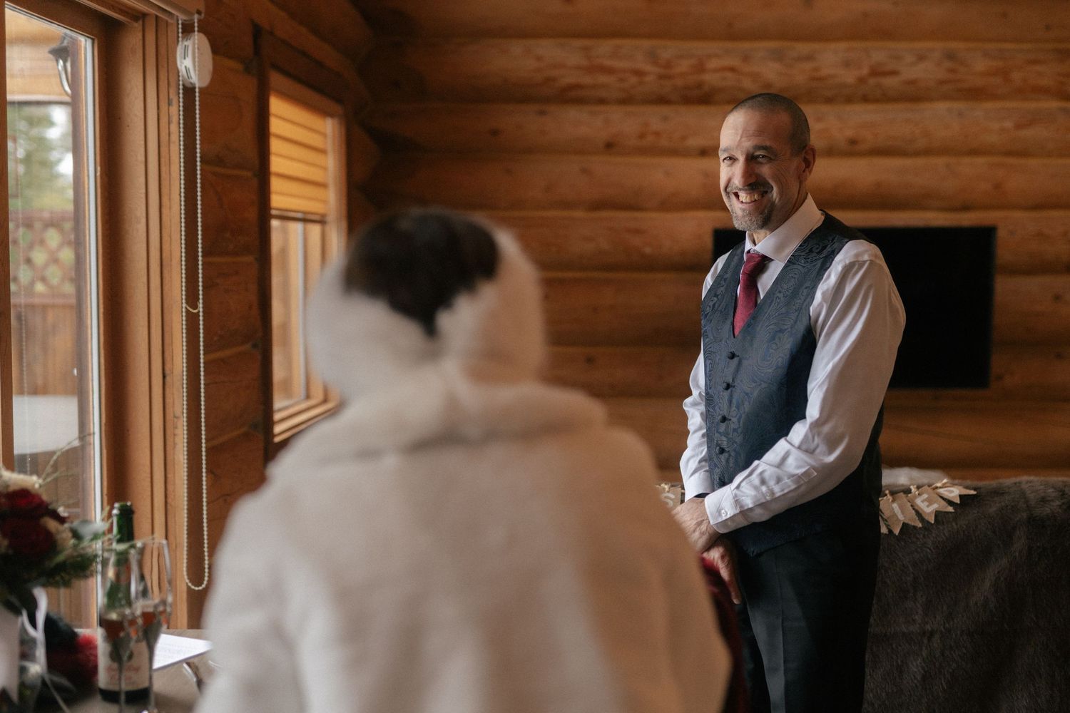 groom grins at bride from across a log cabin