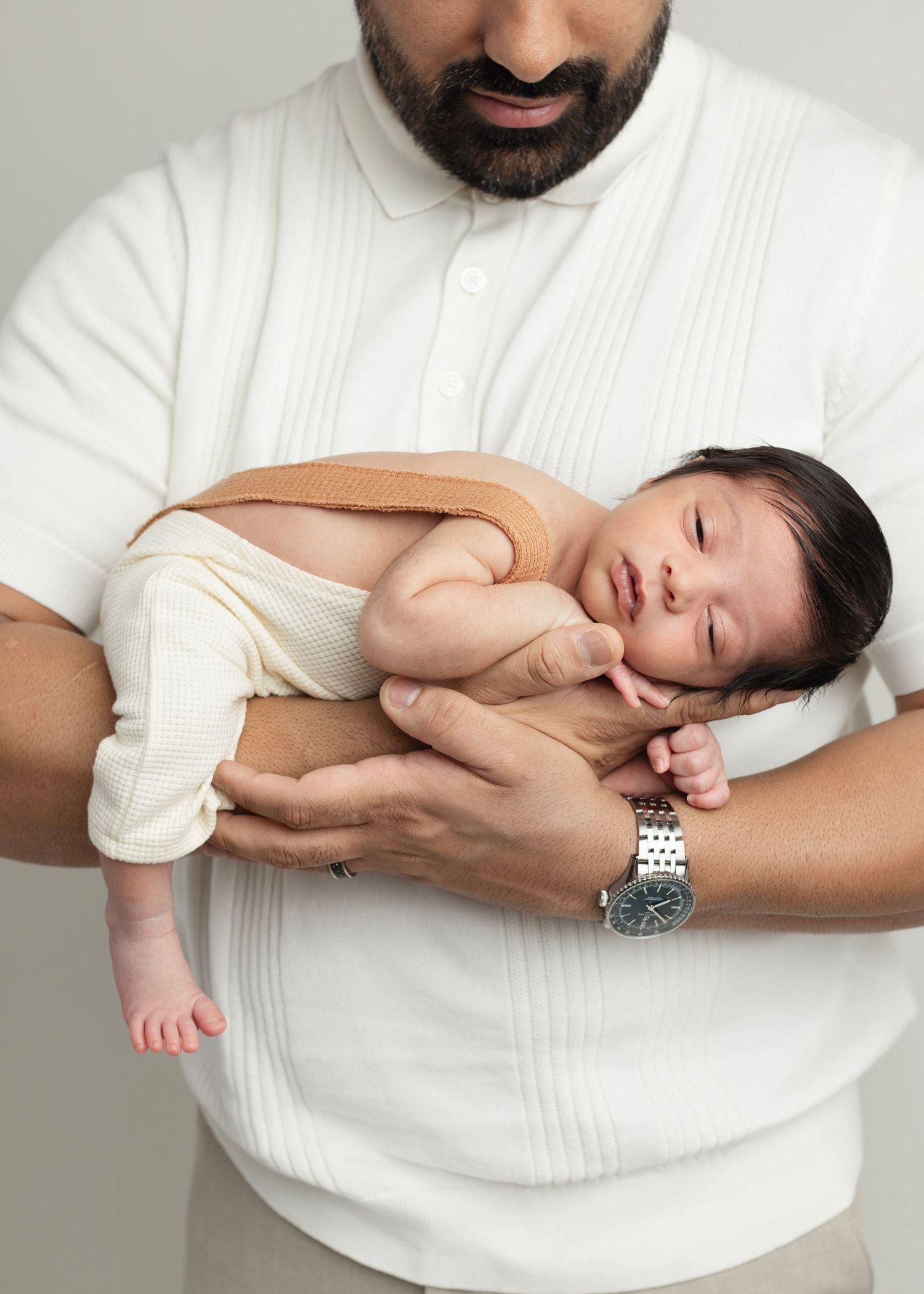 A bearded father in a white polo shirt gently cradles his sleeping newborn baby while wearing a silver watch. Elizabeth Lang Photography Sur