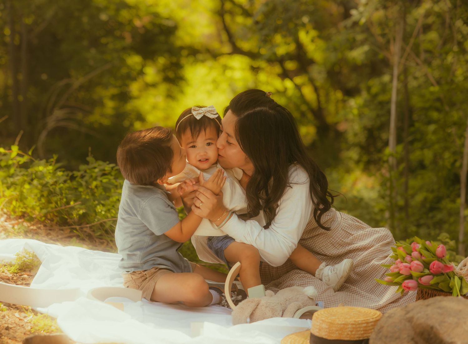 A mother kisses her baby girl while her toddler son sits beside them on a white blanket outdoors photographed by  Yoyo Leung