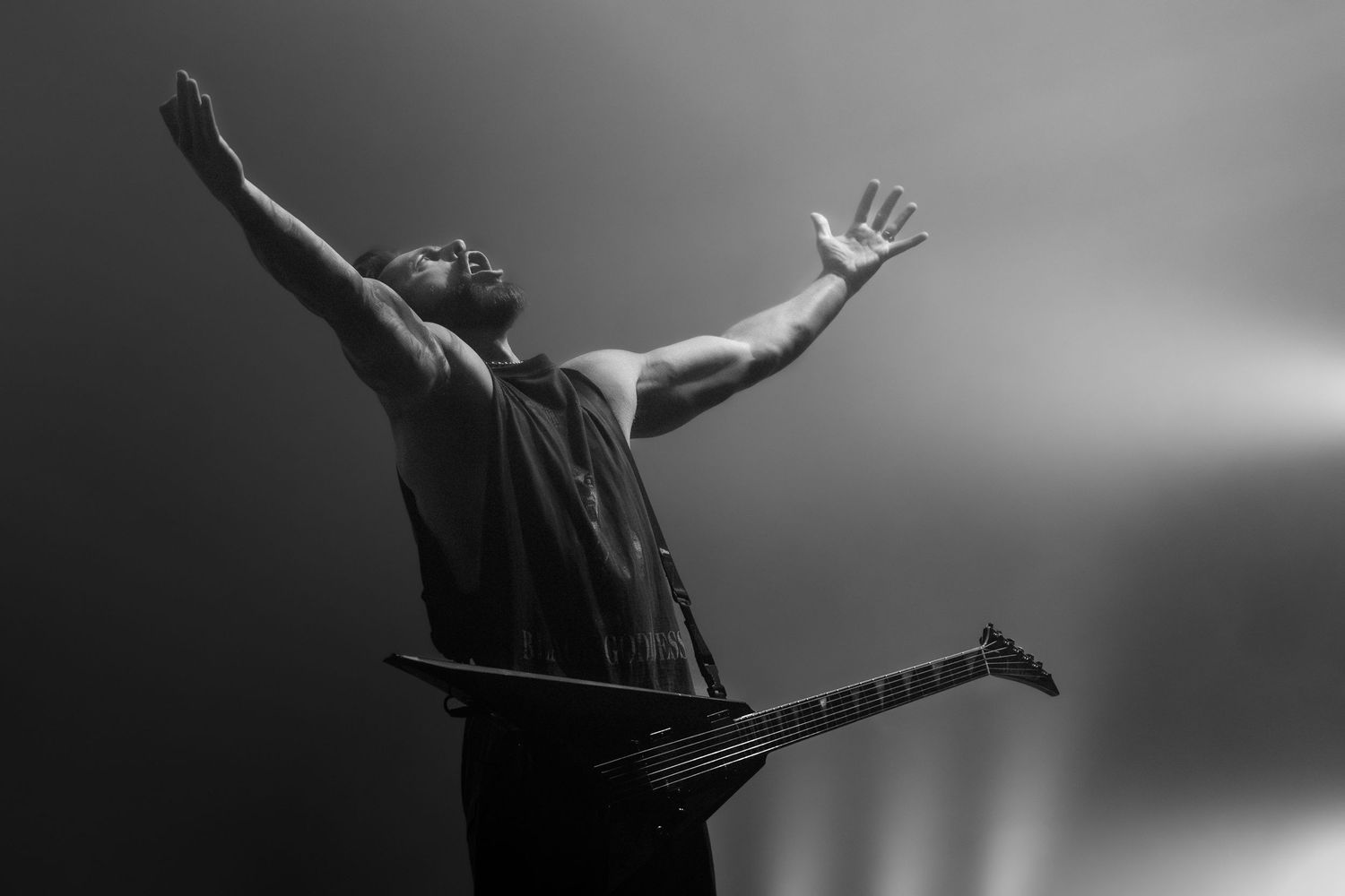 Black and white photo of a guitarist raising arms dramatically on stage with electric guitar, backlit by fog lights.
