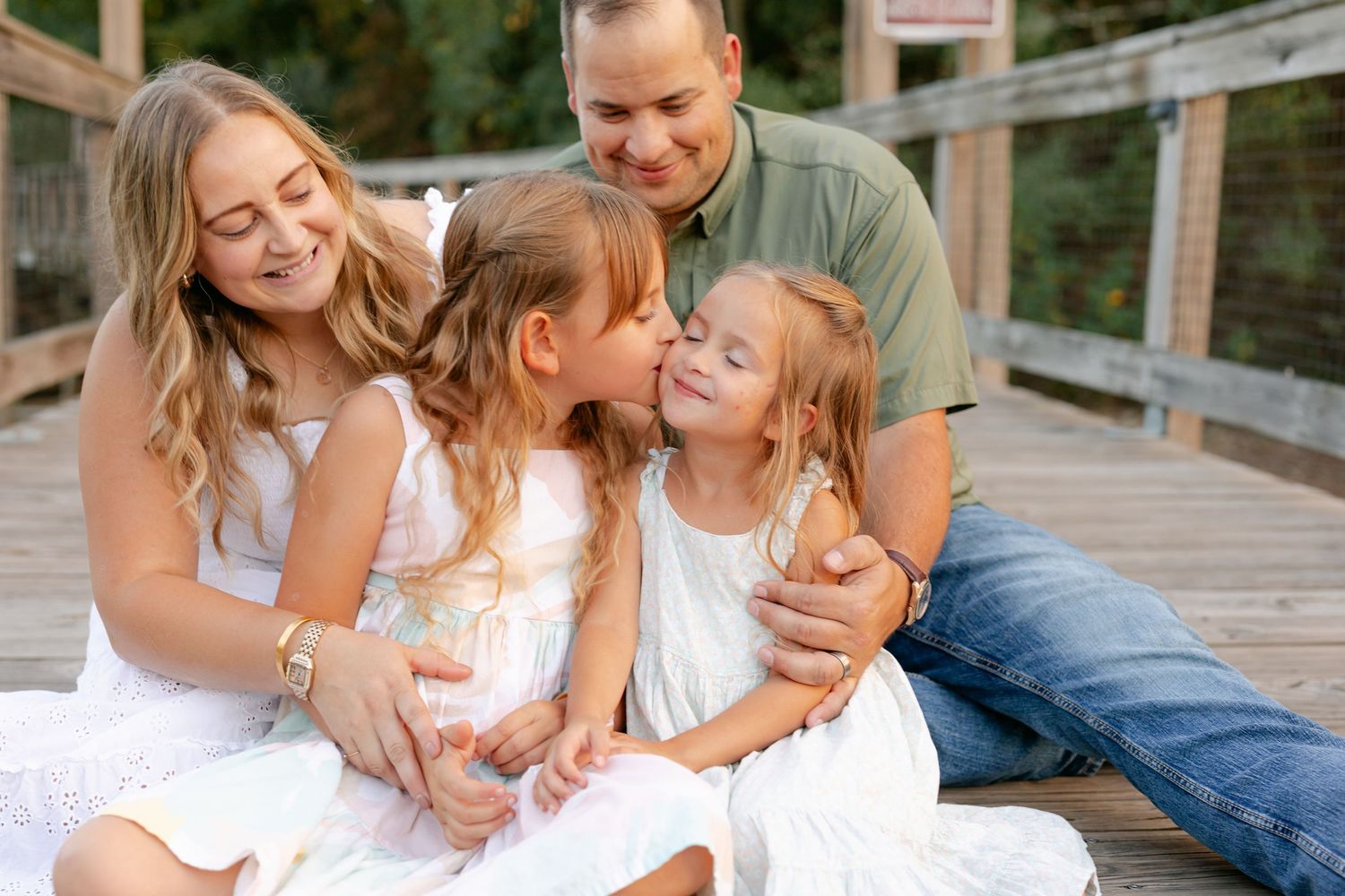 Happy family with two daughters sharing a kiss on a wooden bridge outdoors.