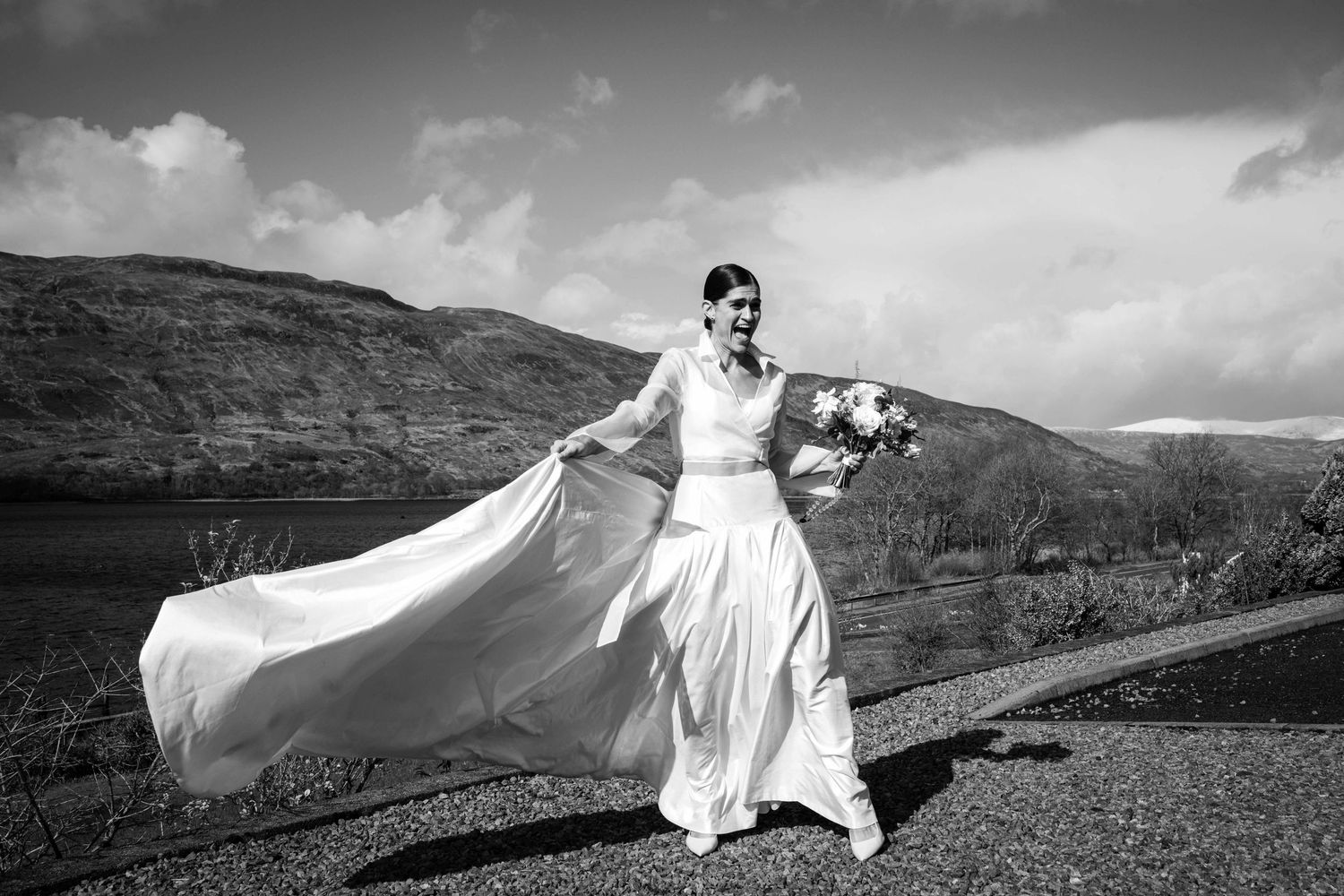 Bride in flowing white wedding gown holding bouquet by a loch with mountains in background, black and white photo.
