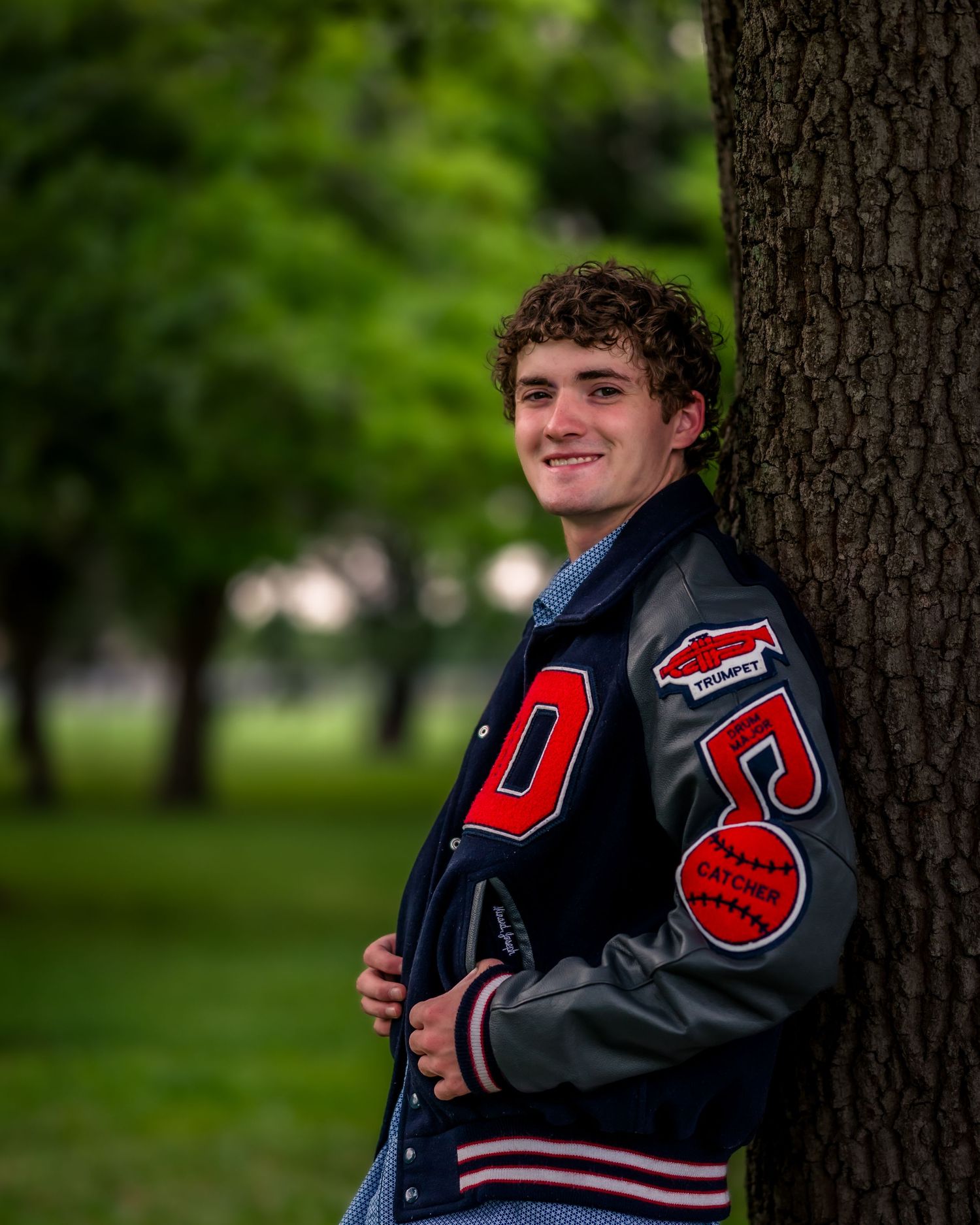 Young man in a varsity letterman jacket leaning against a tree in a green park, smiling for senior photos.