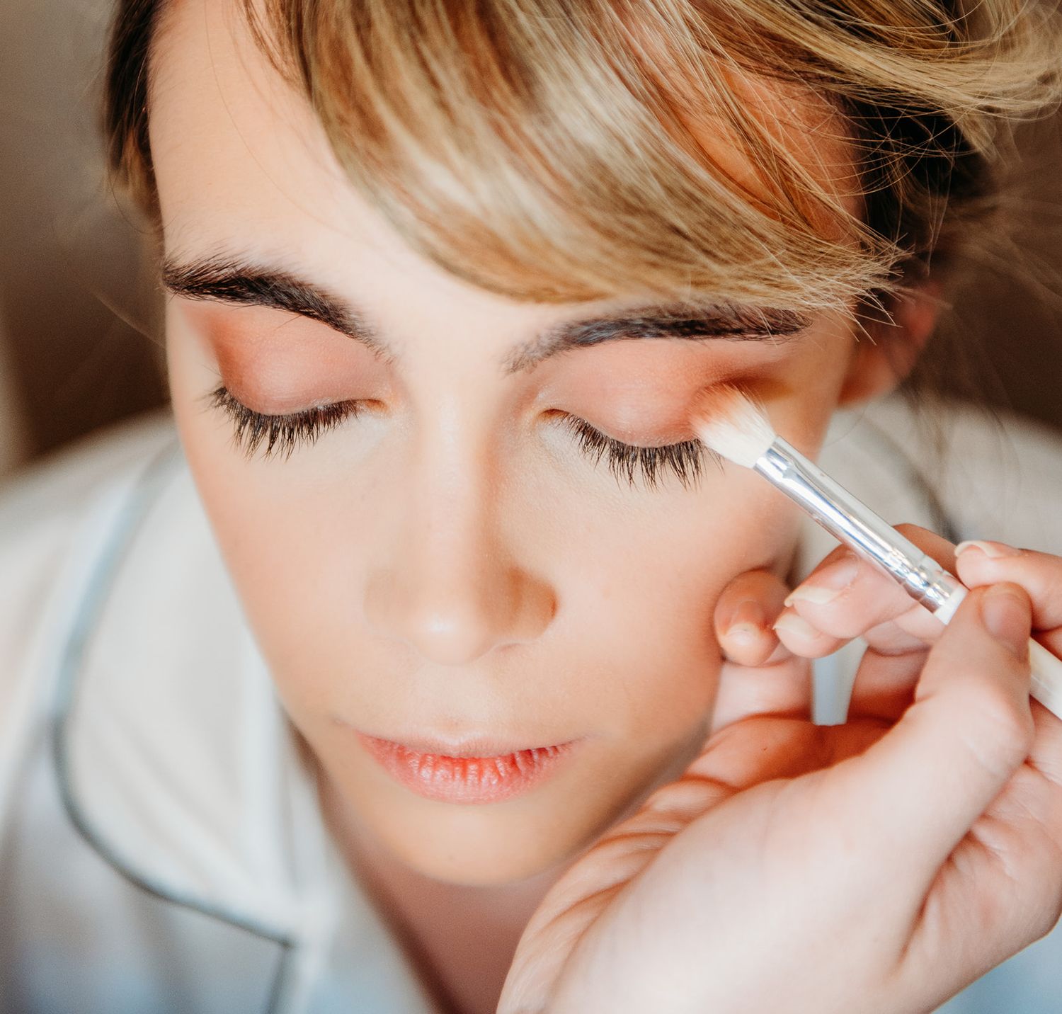 Makeup artist applying eyeshadow with brush to woman with blonde hair and long lashes.