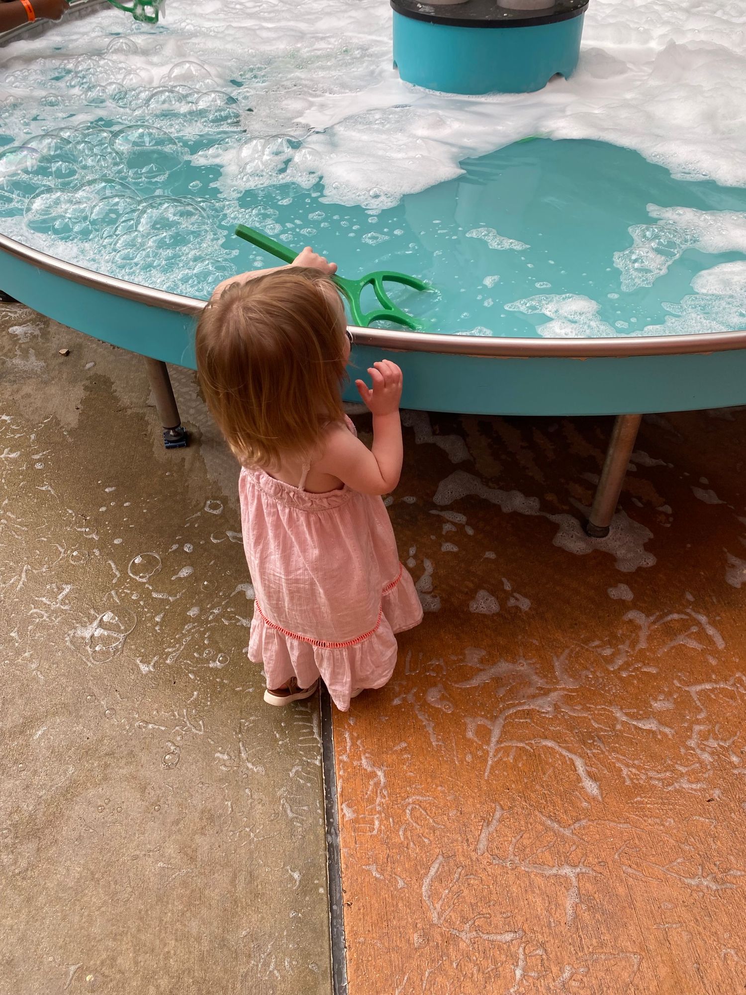Toddler girl in pink dress playing at a water table with foam and green toys at an outdoor play area.