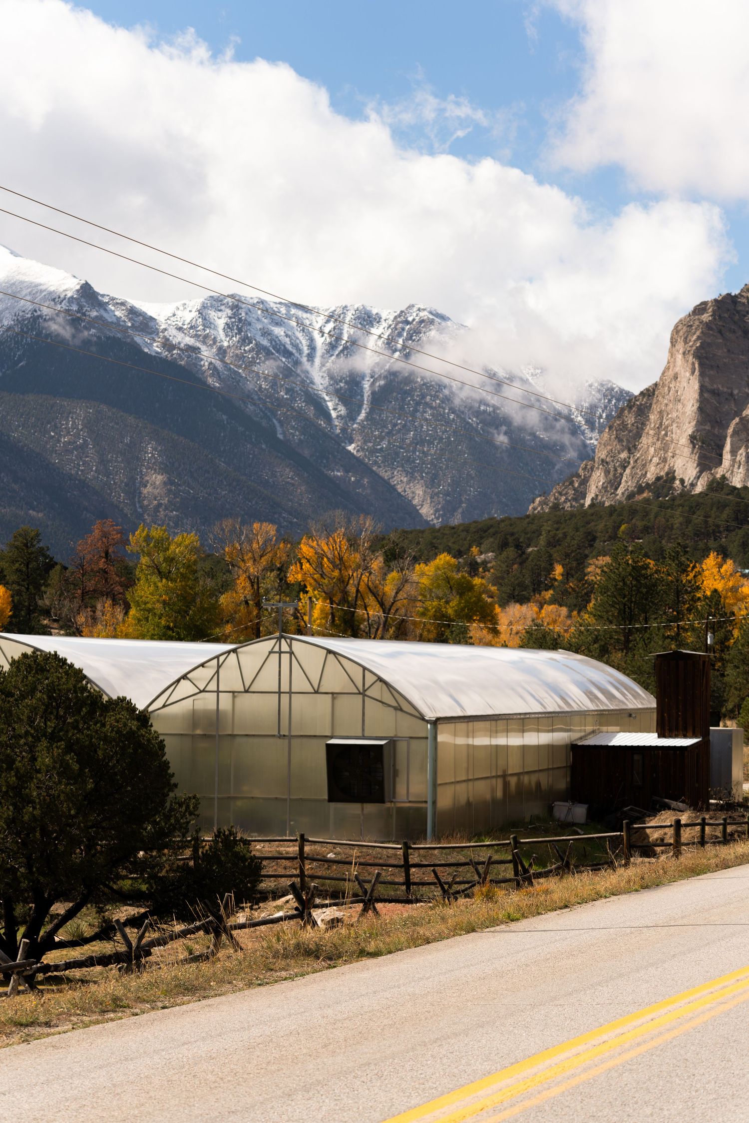 Row of greenhouse structures with snow-capped mountains and autumn trees in the background.