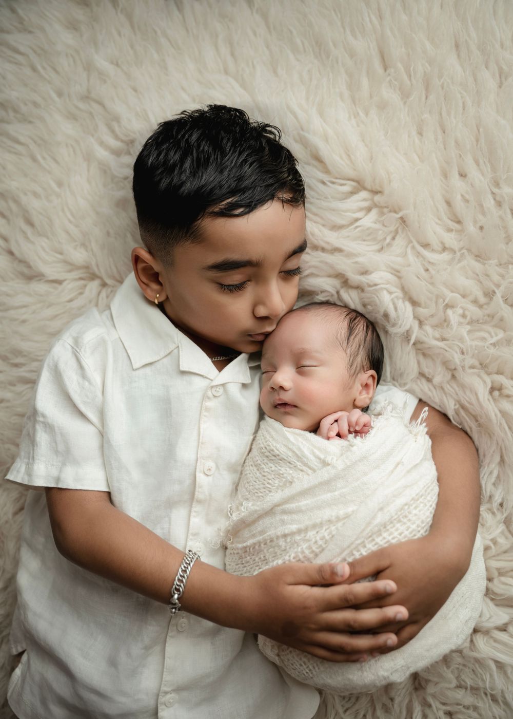 Older boy gently kisses swaddled newborn baby sibling while lying on a soft cream fur backdrop.