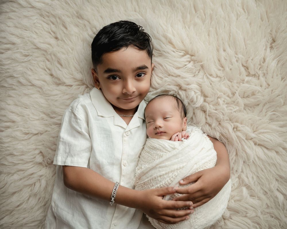 Young boy lying on fluffy rug gently holding swaddled newborn baby sibling in soft white wrap.