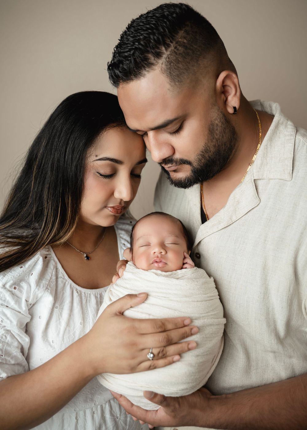 A couple tenderly holds their swaddled newborn baby, sharing an intimate family portrait in a soft, neutral studio setting.