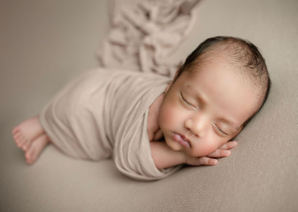 A peacefully sleeping newborn baby swaddled in a soft beige wrap, resting on a neutral gray backdrop.