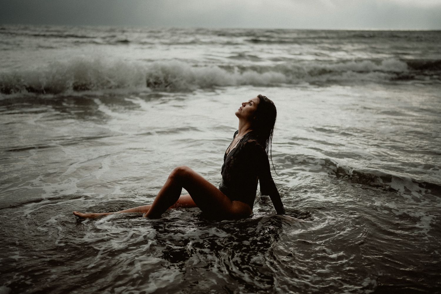 Woman sitting in shallow ocean waves on stormy beach with head tilted back.