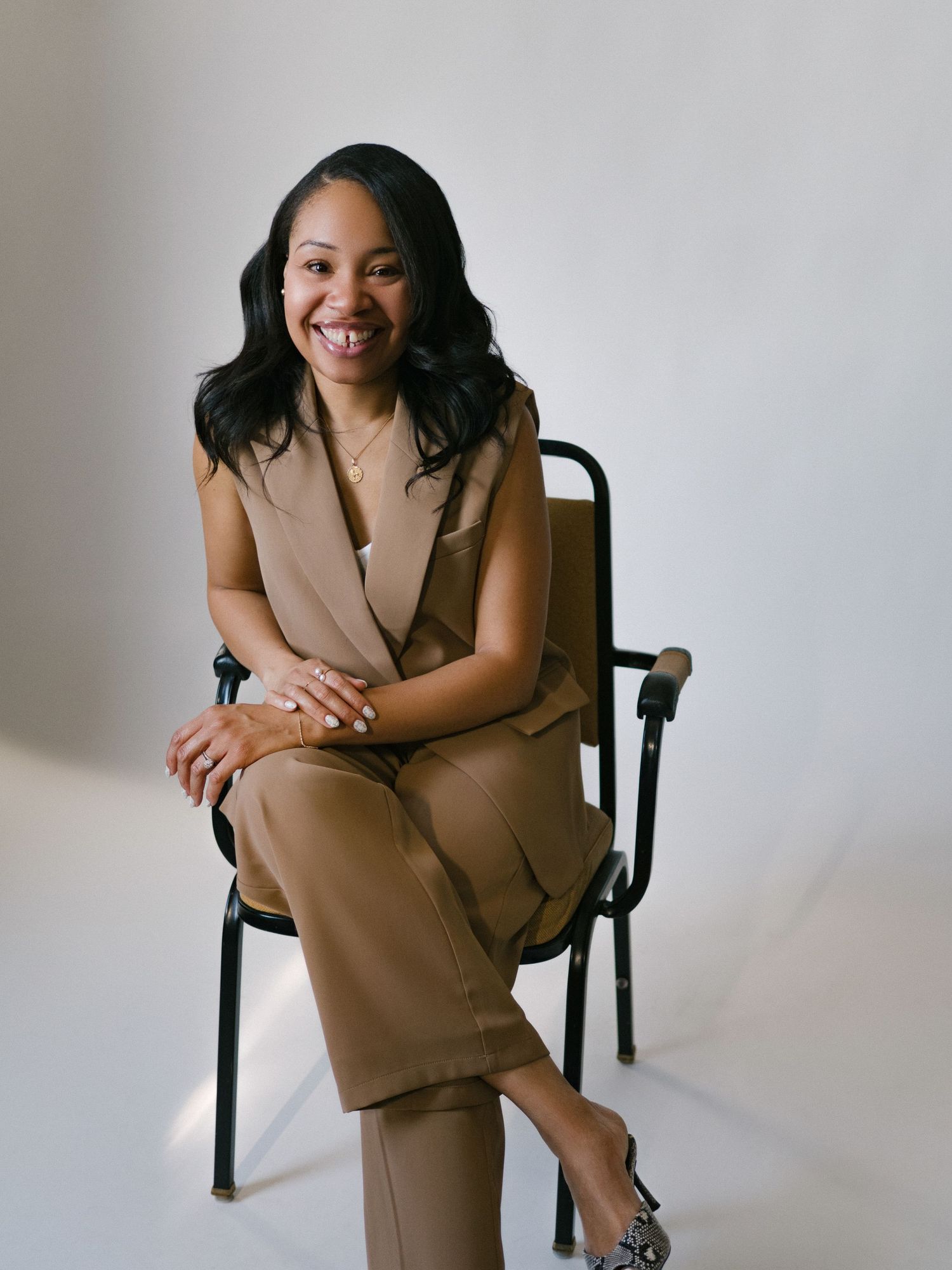 A smiling woman in a tan sleeveless suit sits confidently on a black chair against a white background.