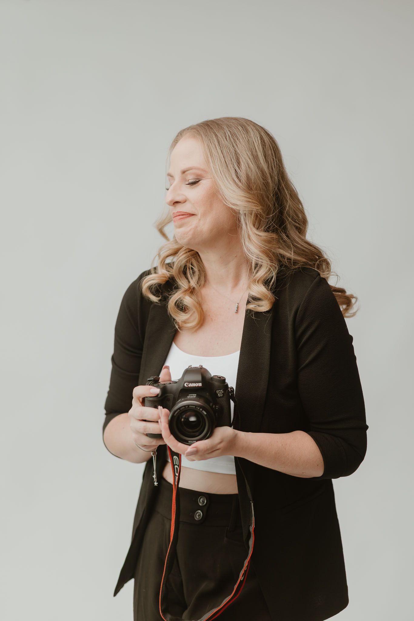 Smiling female photographer in white off-shoulder top holding a DSLR camera against a gray background.