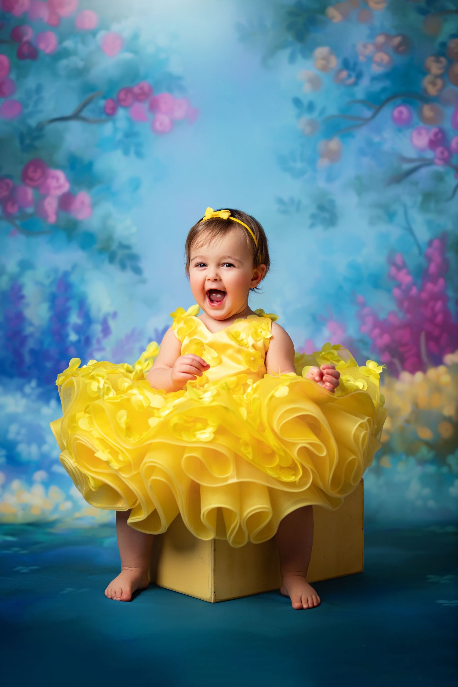 Joyful baby girl in a yellow ruffled dress sits on a yellow box against a colorful floral blue backdrop.