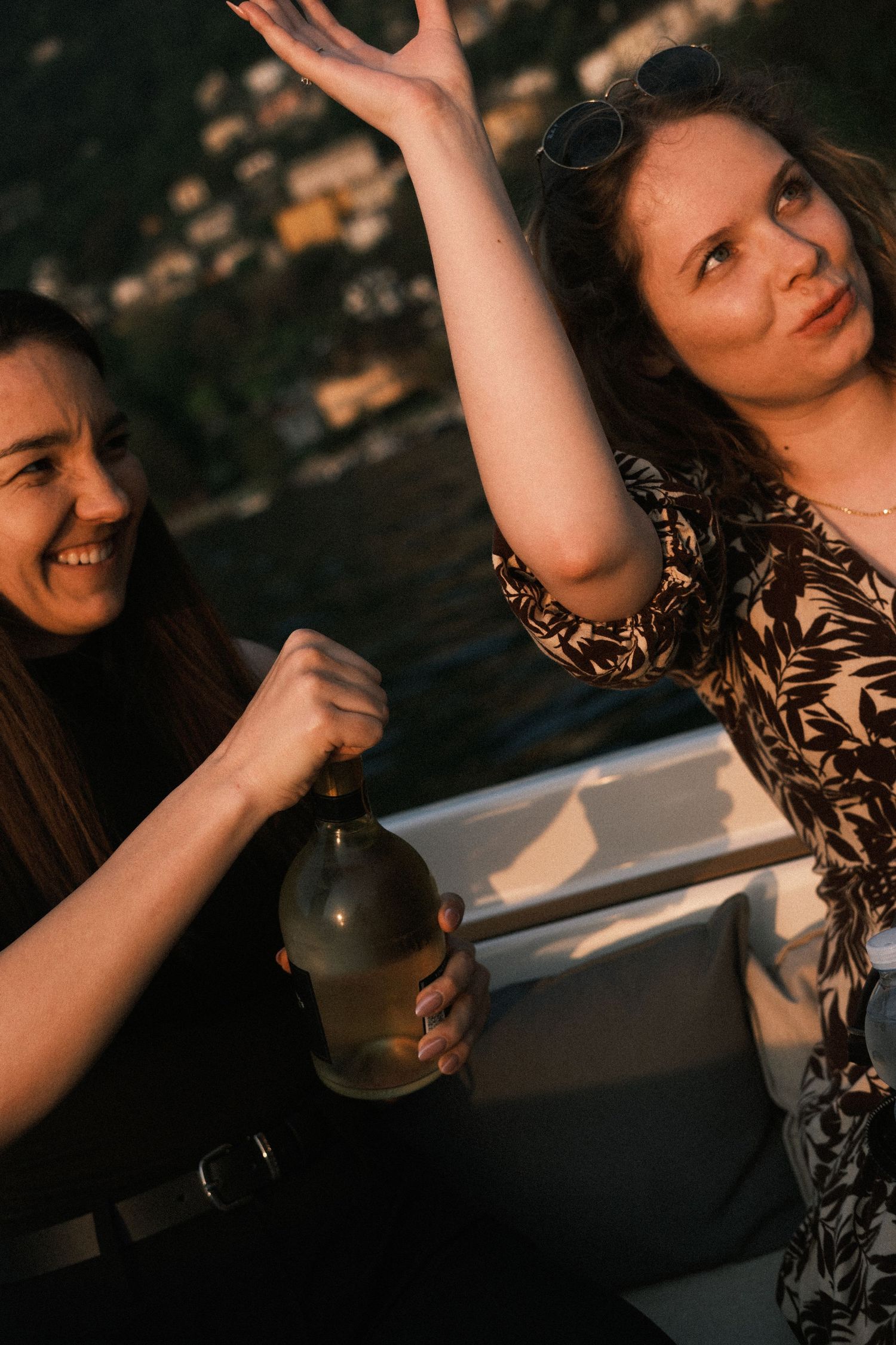 Two women celebrating outdoors on a boat at sunset, one holding a bottle while the other raises her hand.