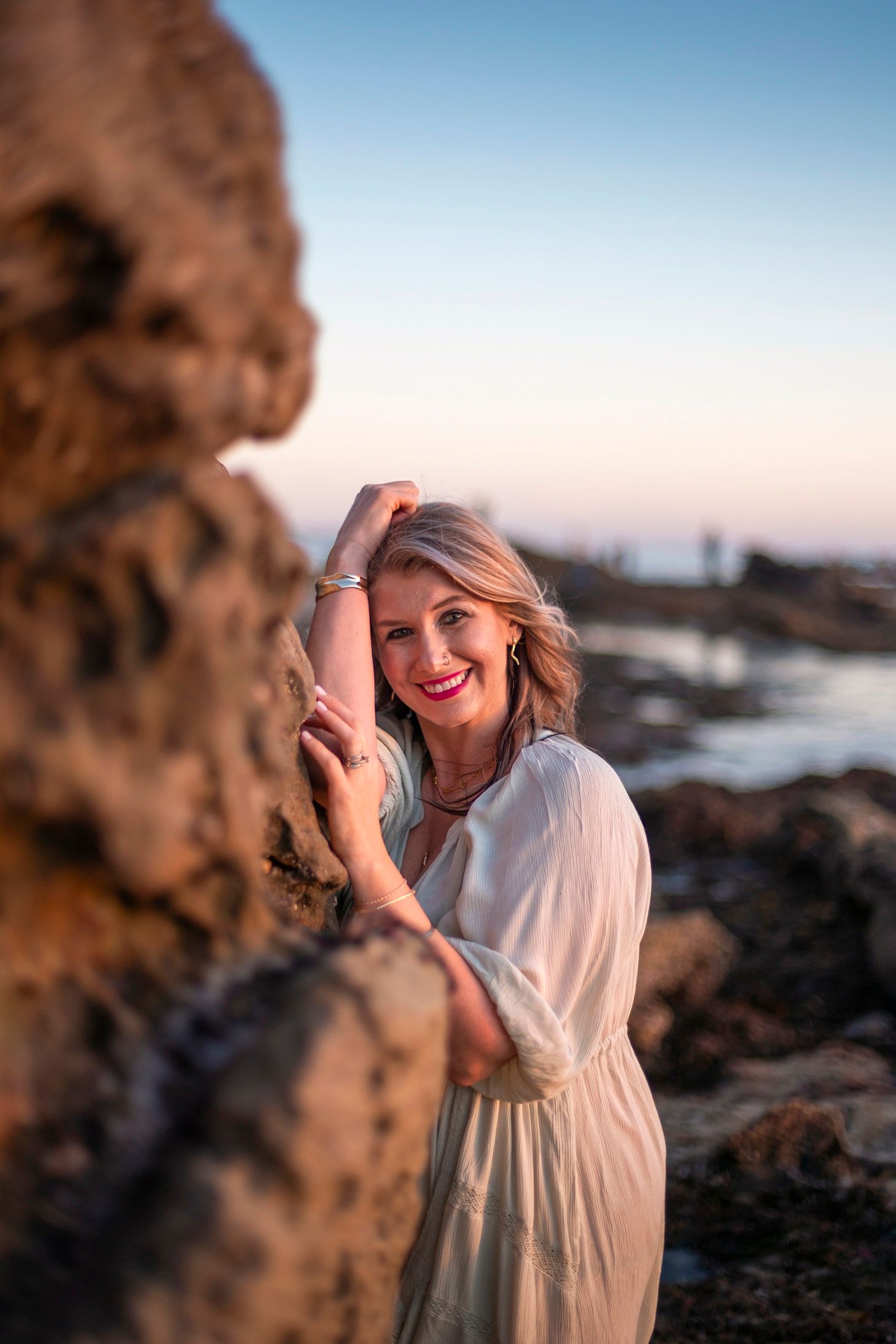 Blonde woman in white dress smiling beside rocky coastal formation at sunset by the ocean.