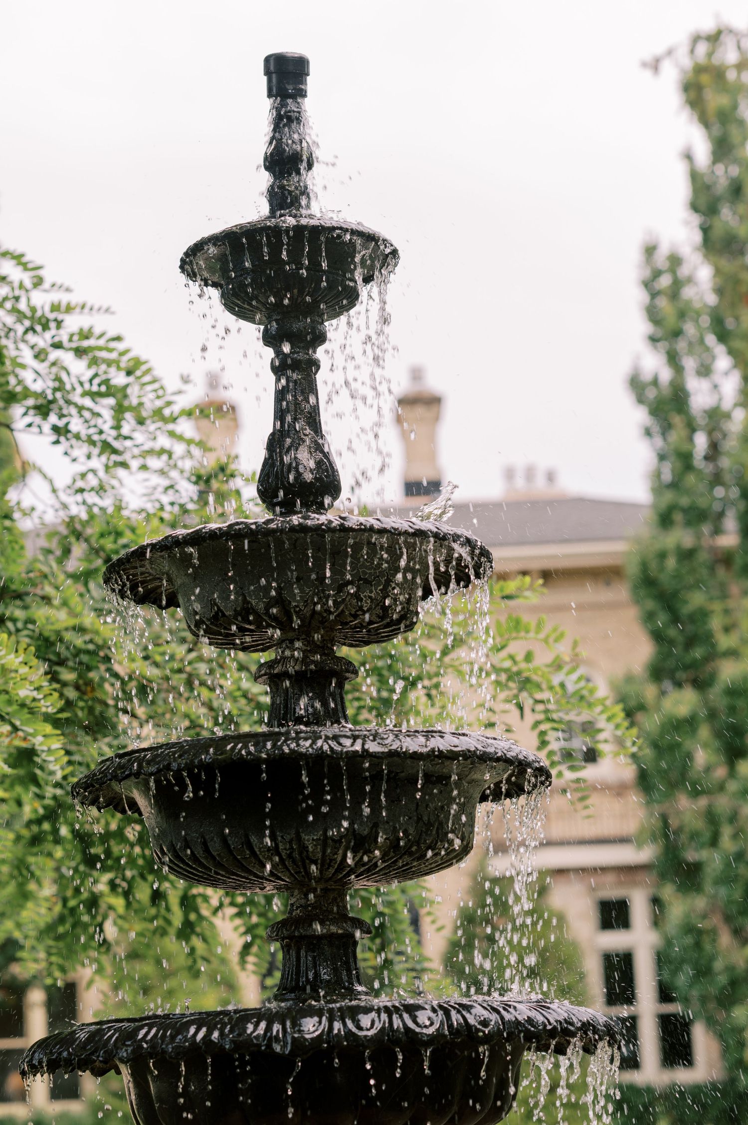 A classic three-tiered black cast iron garden fountain with cascading water, surrounded by lush green trees and a stone building.
