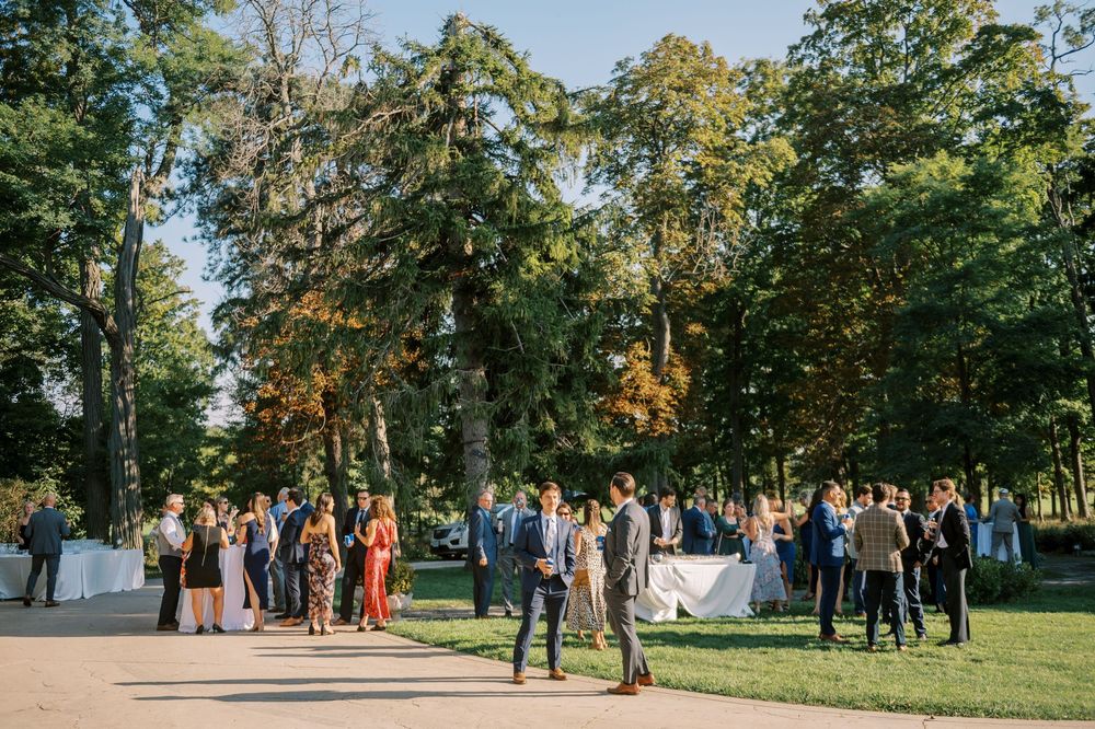 Elegant outdoor wedding cocktail hour with guests mingling on a tree-lined path in a lush garden setting.