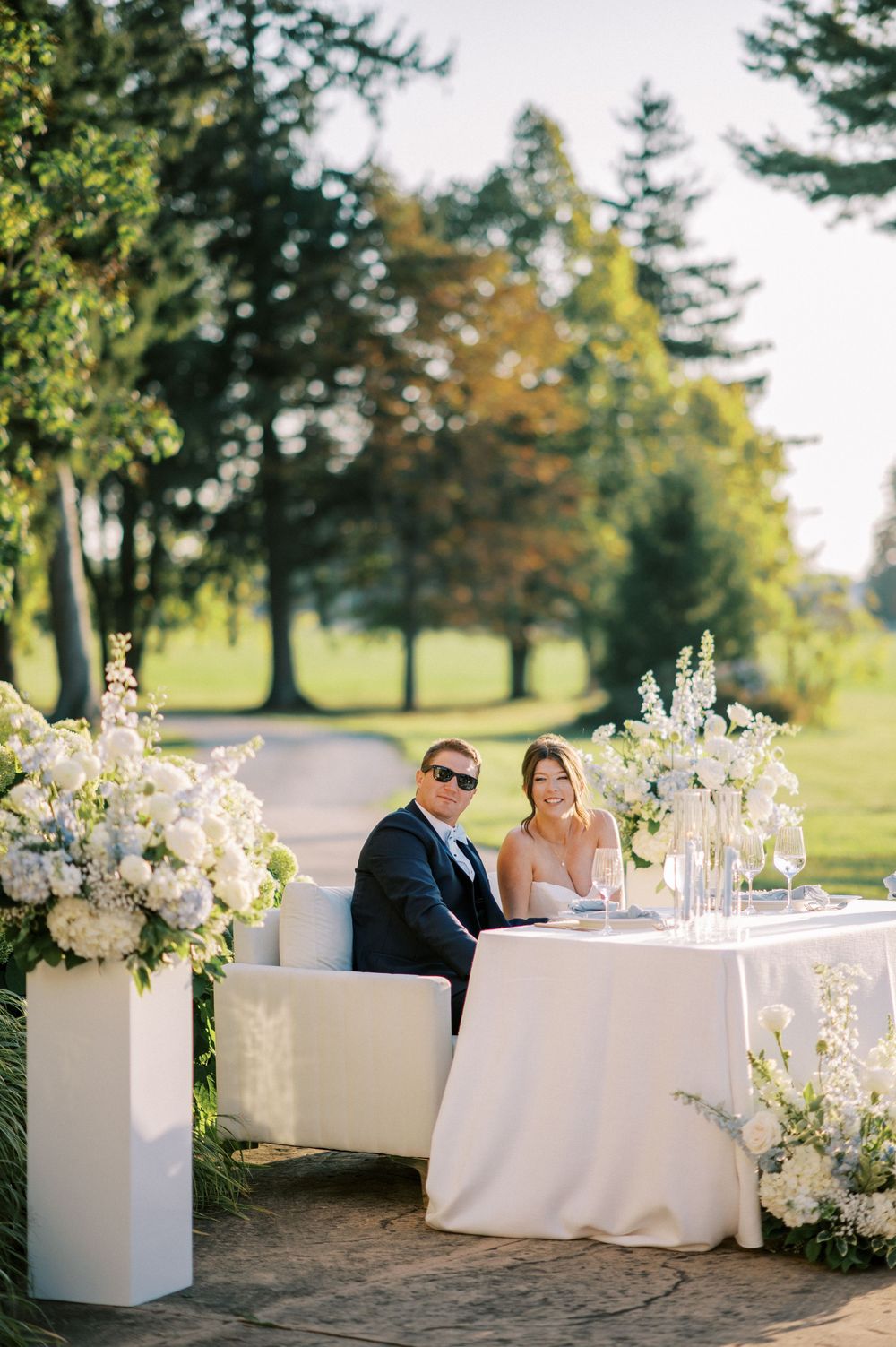 Bride and groom seated at elegant white sweetheart table with tall floral arrangements outdoors.
