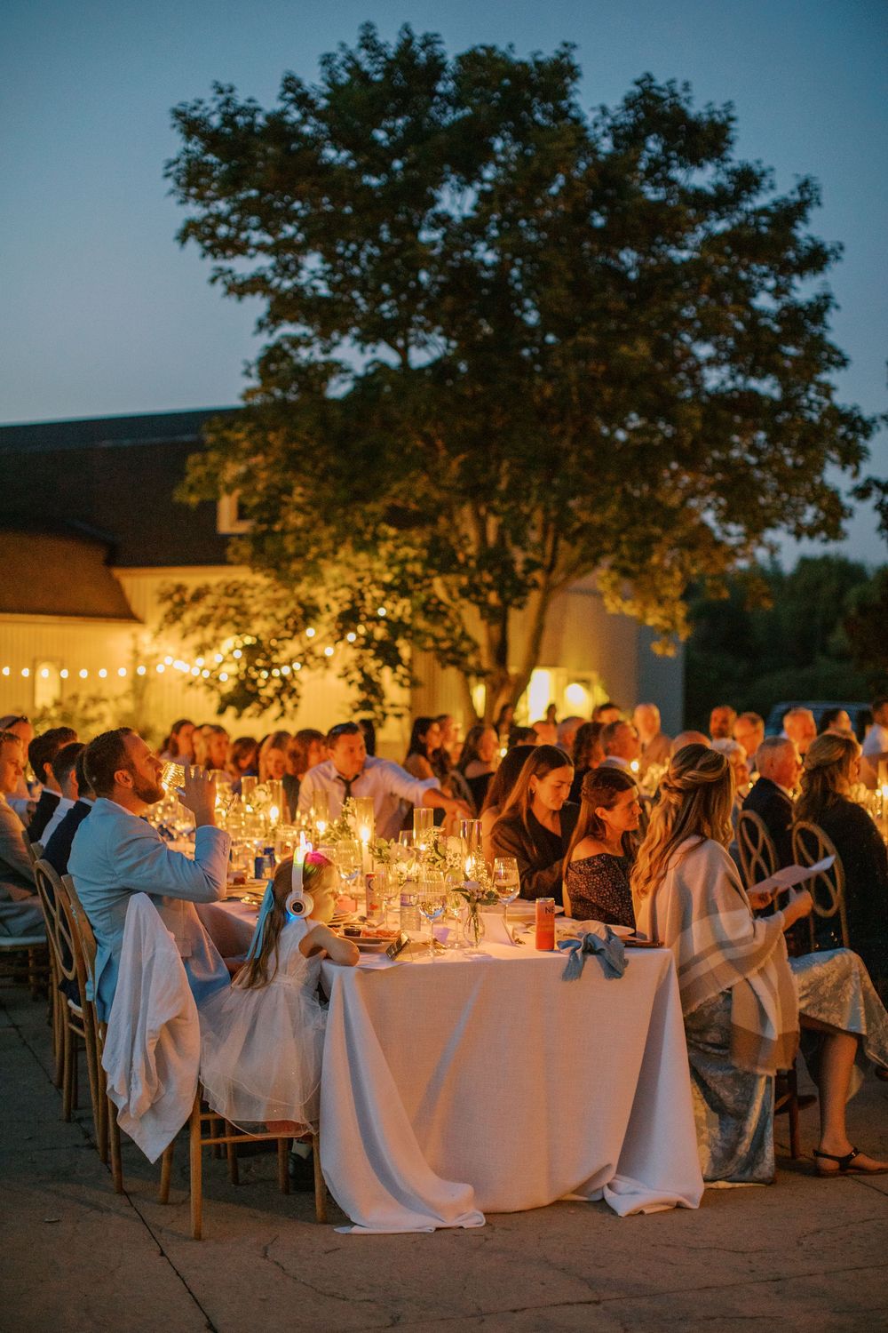 Elegant outdoor wedding reception at dusk with candlelit tables, string lights, and guests dining under a large tree.