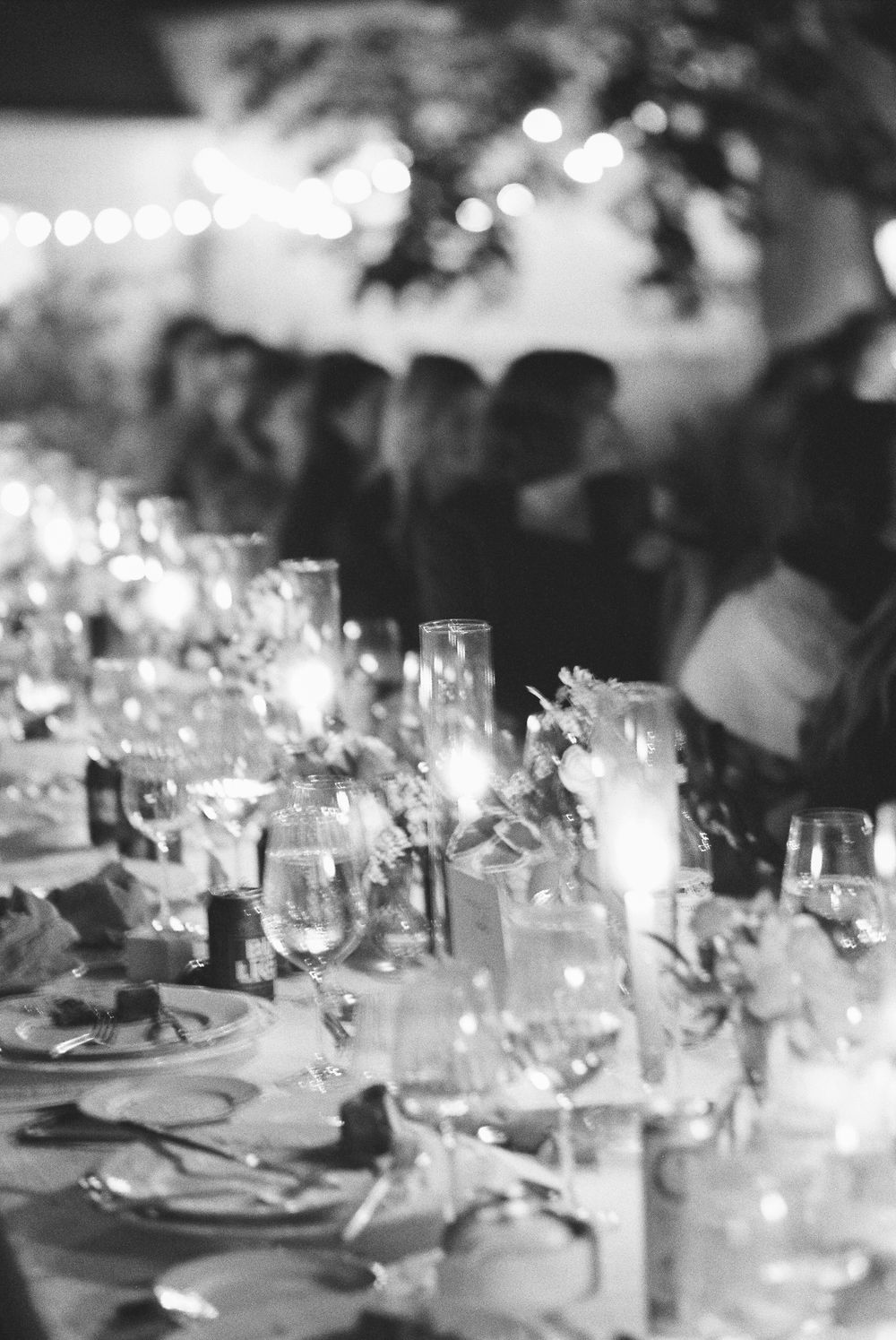 Black and white wedding reception table with elegant glassware, candles, and floral arrangements with guests in background.