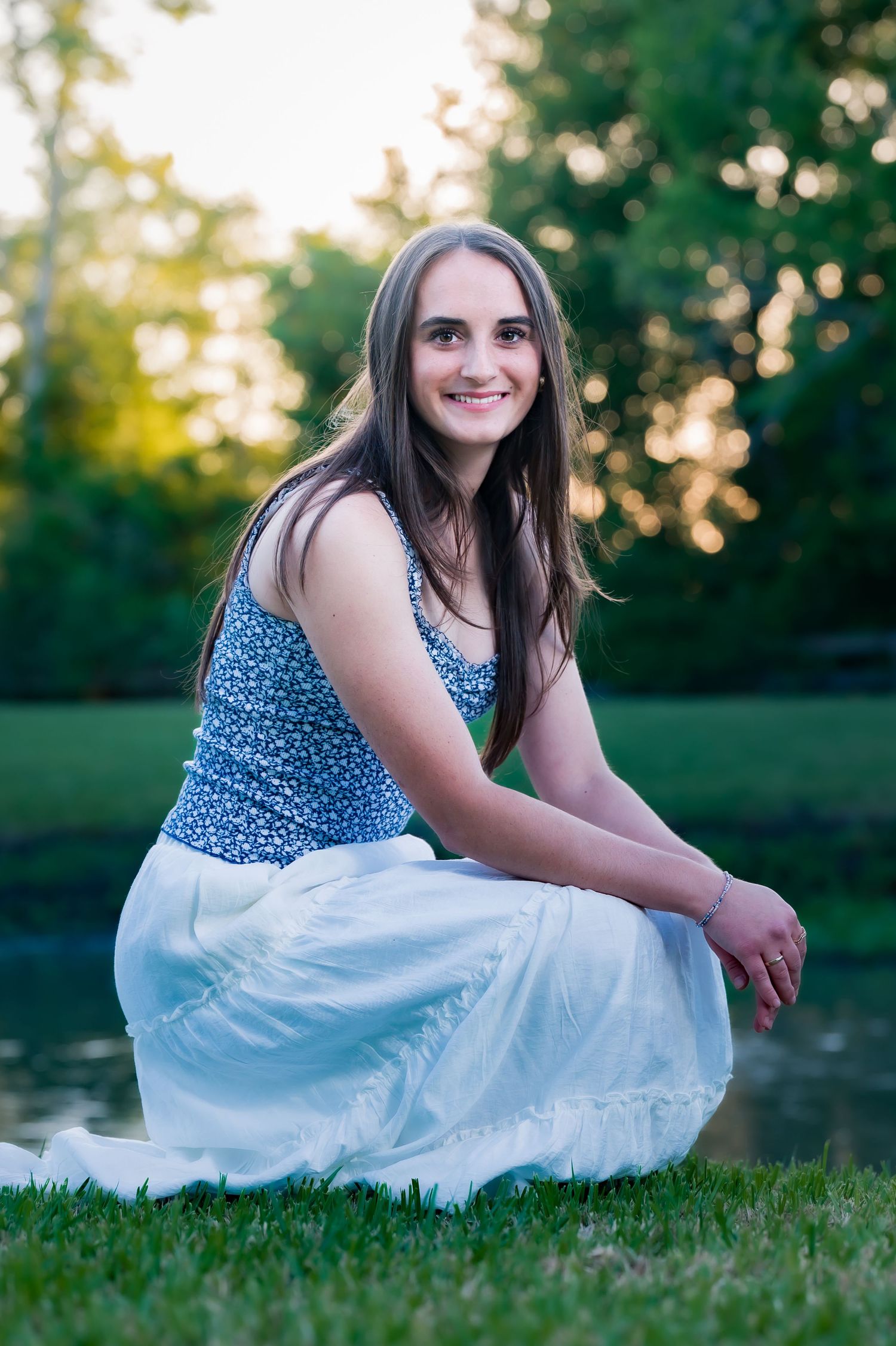 Young woman in burgundy dress holding green shoes smiles by a serene pond surrounded by lush trees.