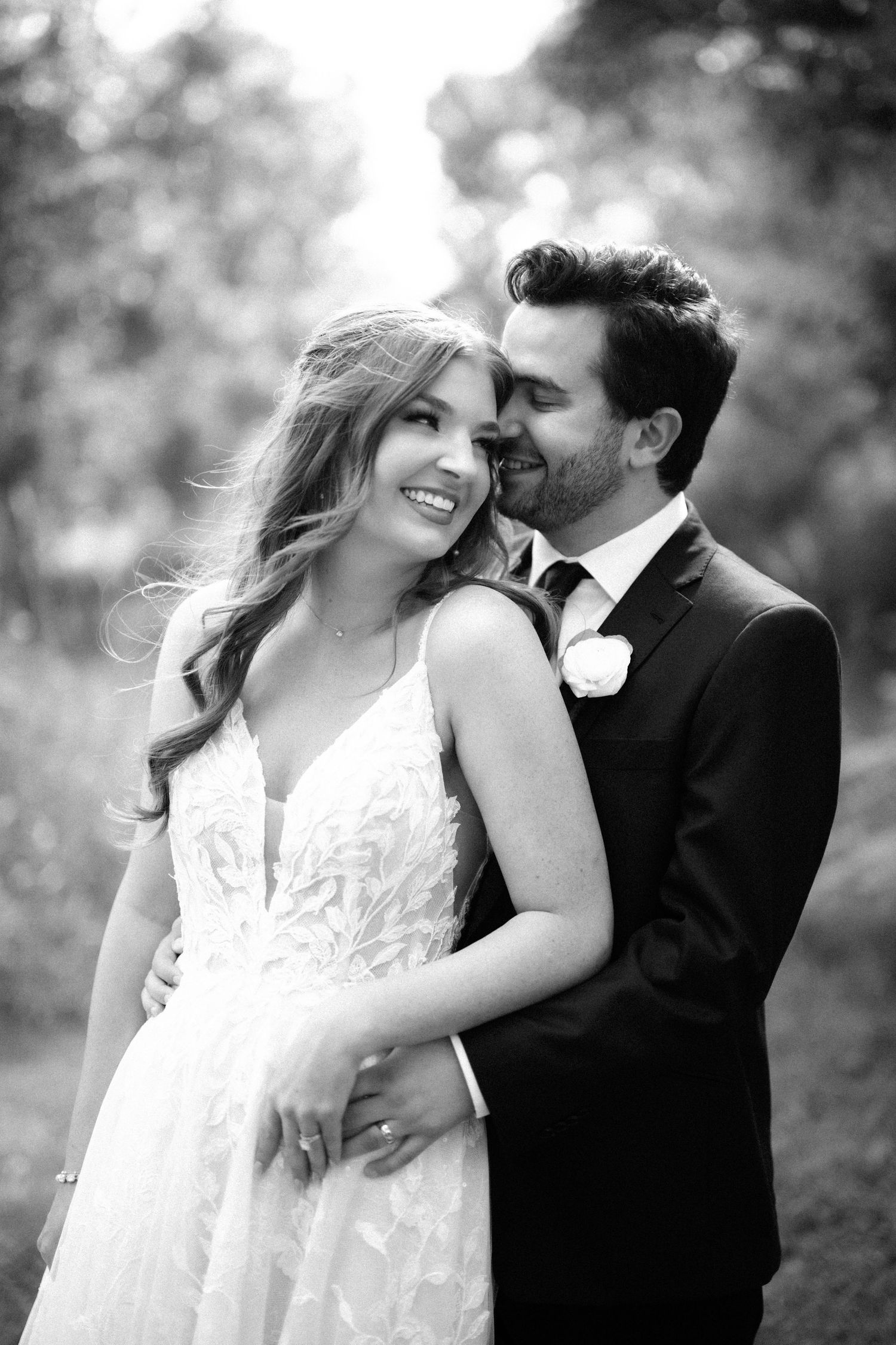 Black and white wedding portrait of smiling bride in lace gown with groom in suit embracing outdoors.