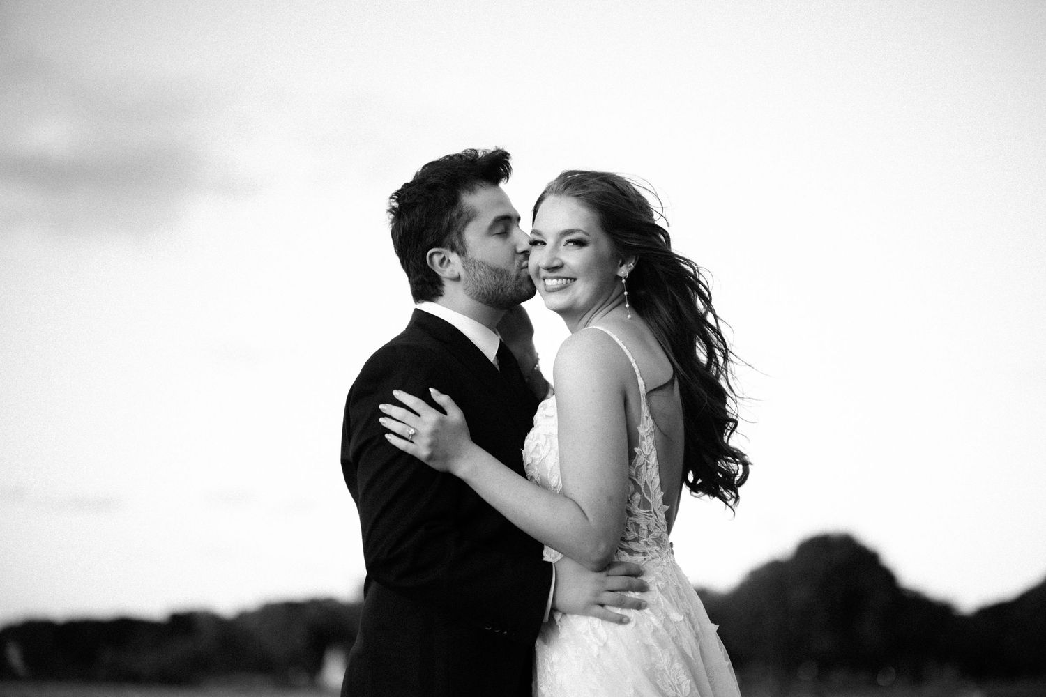 Black and white wedding photo of a couple embracing outdoors, bride in gown smiling, groom in suit at sunset.