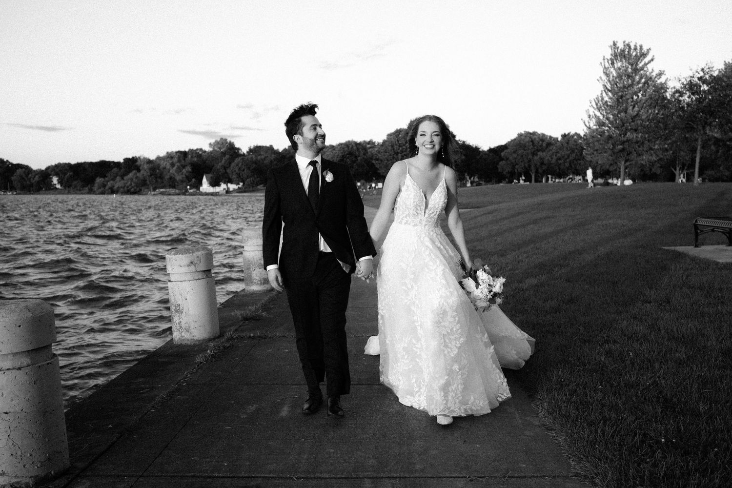 Black and white photo of a bride and groom holding hands, walking along a waterfront path on their wedding day.
