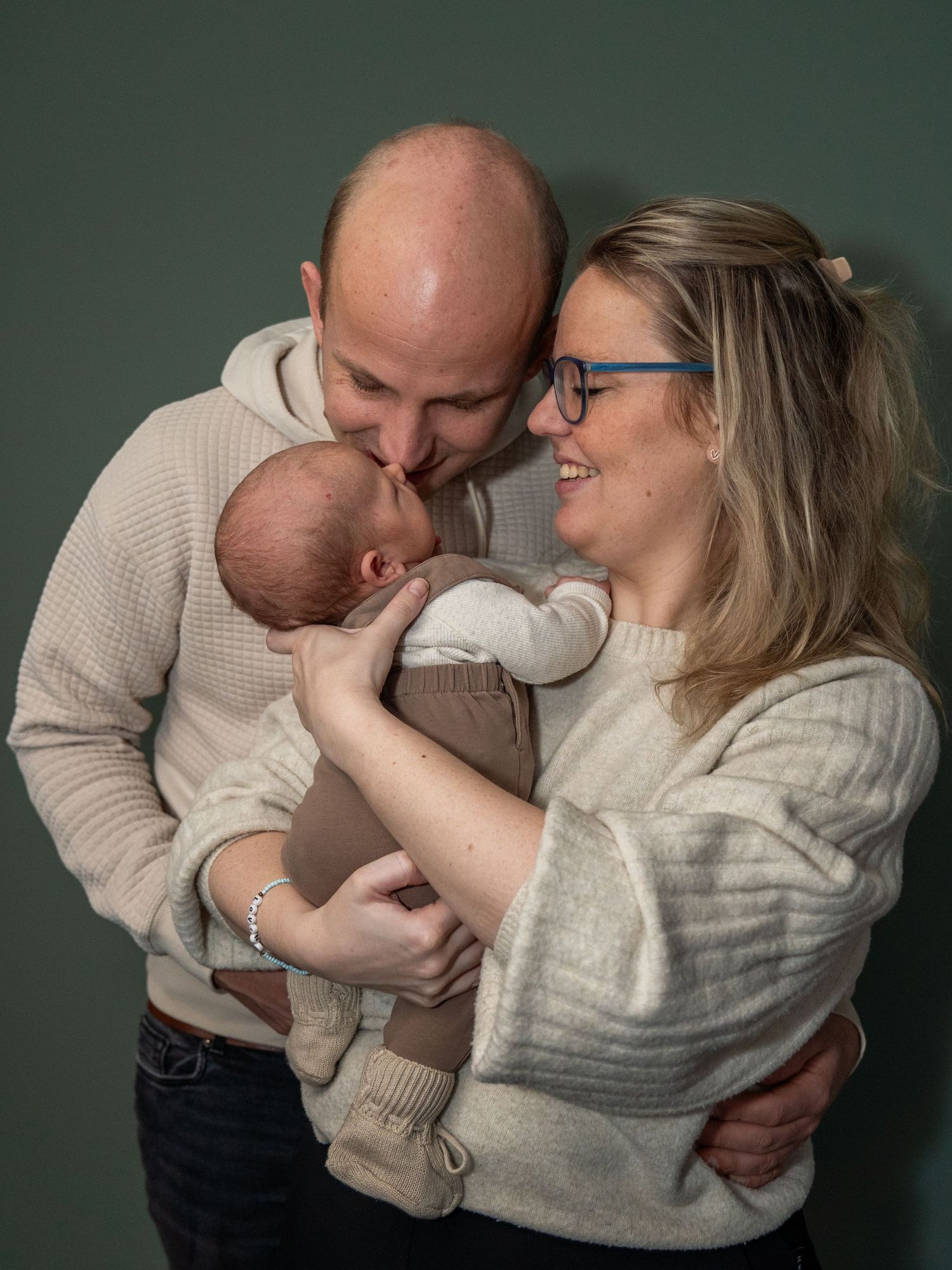 Gelukkige ouders met pasgeboren baby tegen een groene achtergrond; de vader kust het hoofdje van de baby.