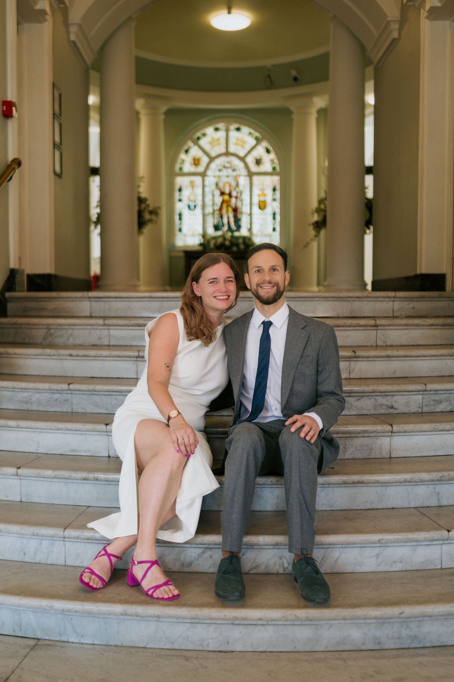 Couple in wedding attire sitting on marble steps inside elegant venue with stained glass window backdrop.