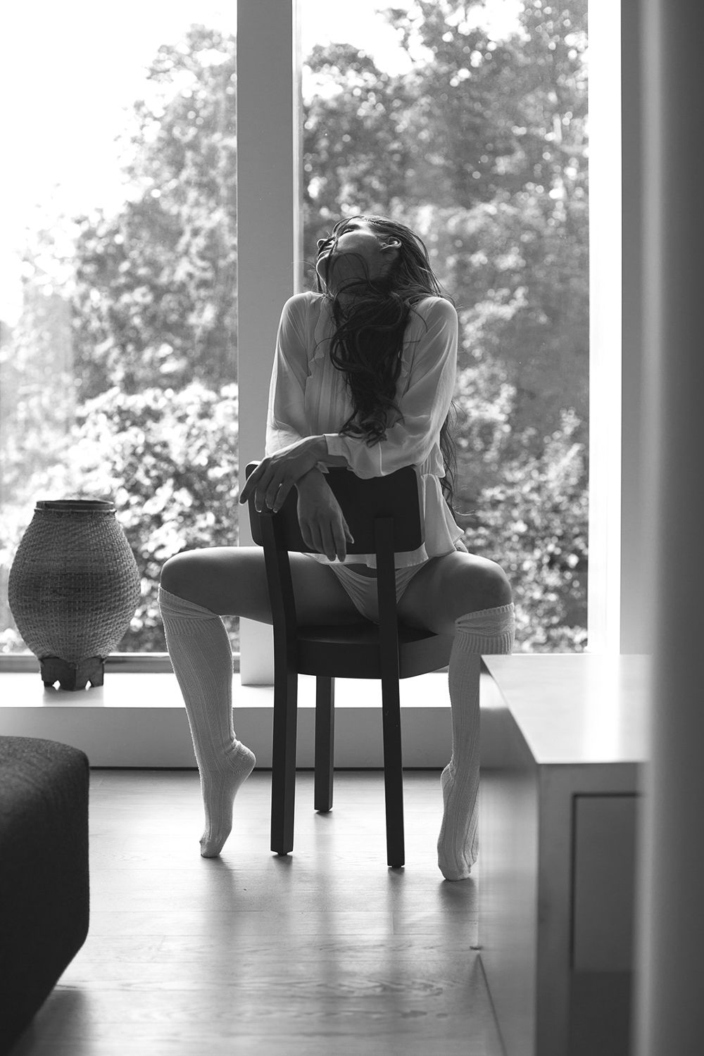 Black and white silhouette of a woman sitting on a chair by a large window with trees outside.