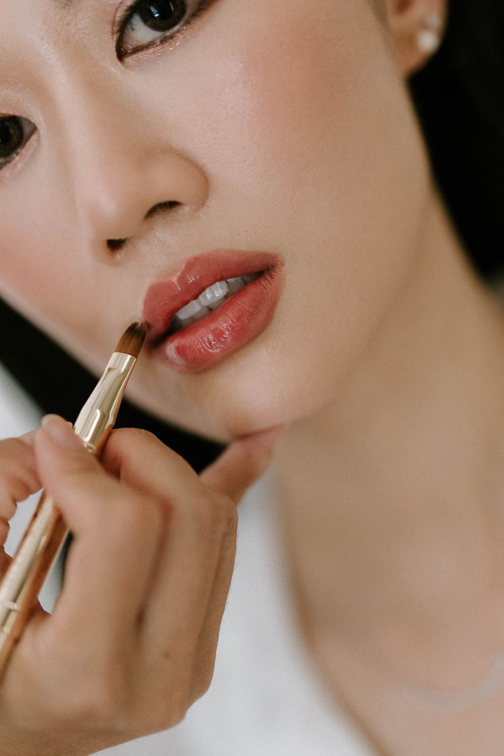 A makeup artist applies rich rose-brown lipstick to a young Asian woman using a professional lip brush.
