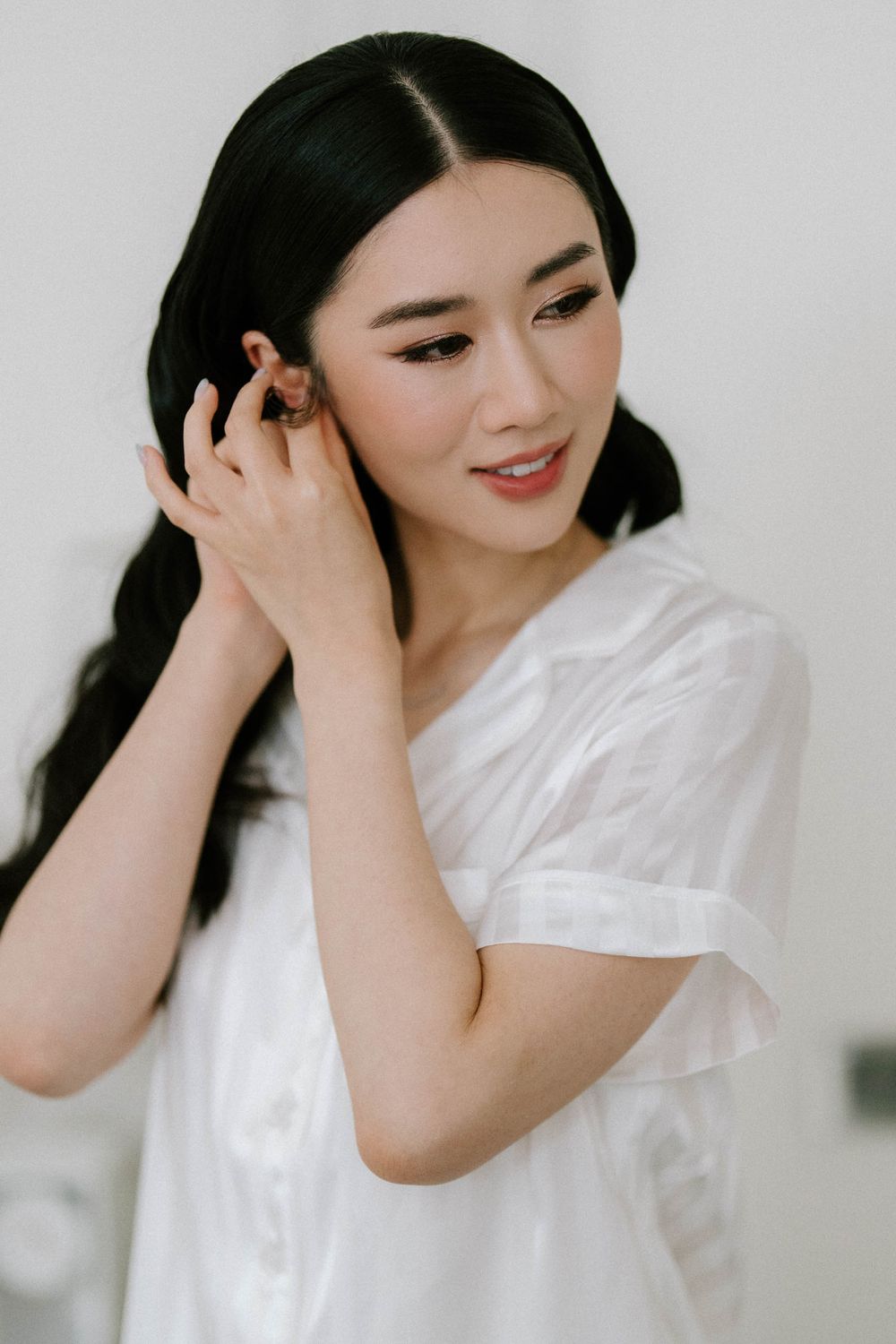Young Asian woman in white dress touching her earring, smiling softly with elegant makeup and dark wavy hair.