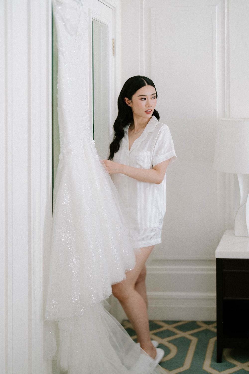 Bride in white satin pajamas holding her lace wedding gown in an elegant hotel room.