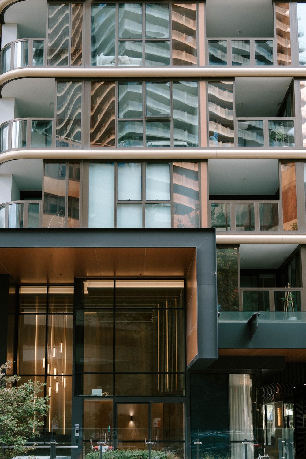Modern luxury apartment building facade with glass balconies, bronze accents, and sleek dark entrance lobby.