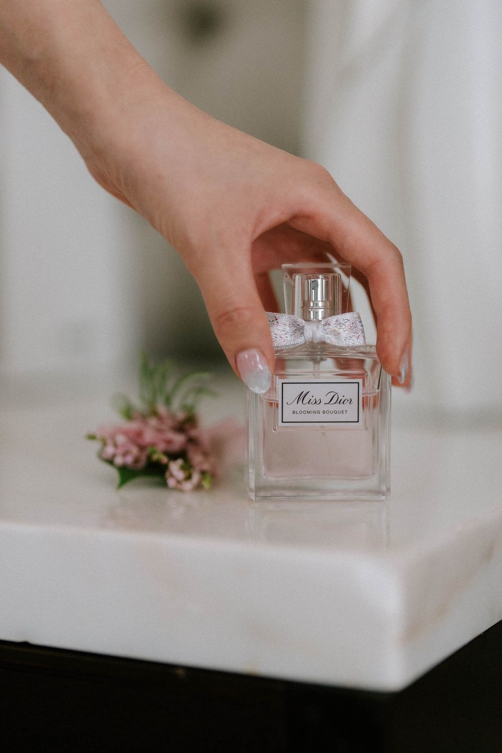 A hand with manicured nails picks up a Miss Dior Blooming Bouquet perfume bottle with a white bow on a marble surface.