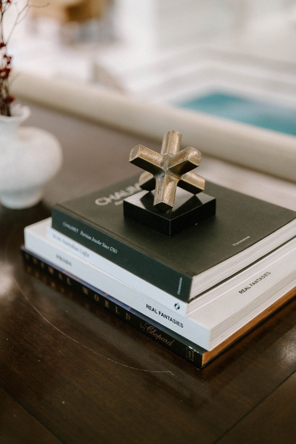 Stack of luxury coffee table books topped with a decorative metal cross sculpture on a dark wooden table.