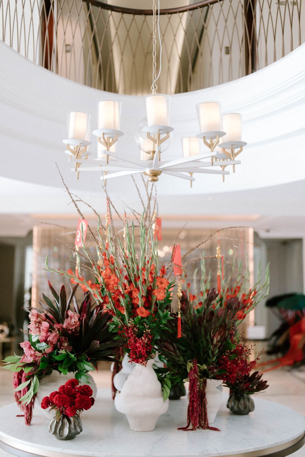 Elegant floral arrangements with red blooms and branches on a white table beneath a chandelier in a bright hotel lobby.