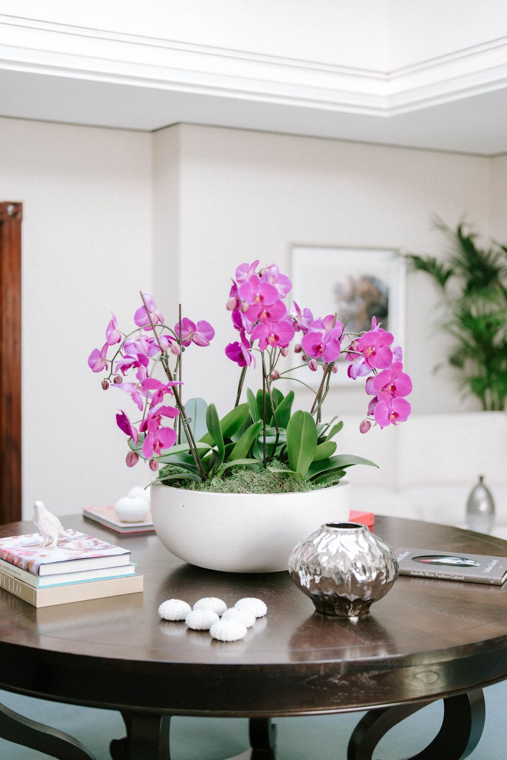 Pink orchids in white bowl centerpiece on dark wood table with books and silver vase in elegant living room.
